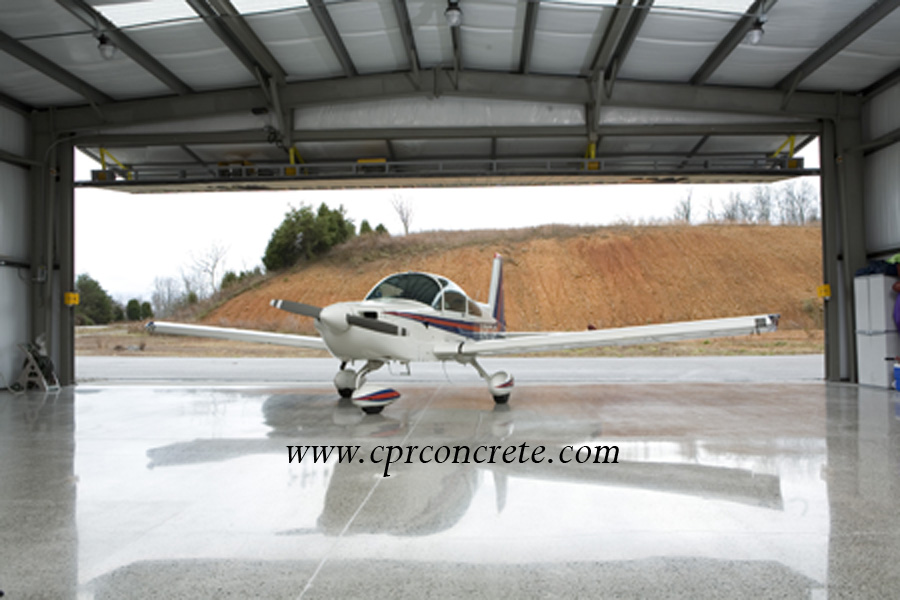 A propeller plane sits on a polished cement floor, facing away from an open hangar door.