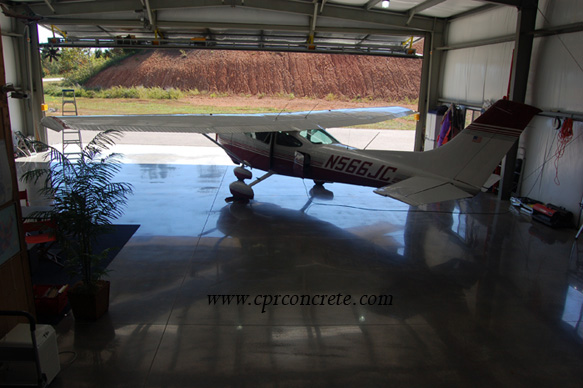 A plane sits on the polished concrete floor of a hangar, facing towards an open door.