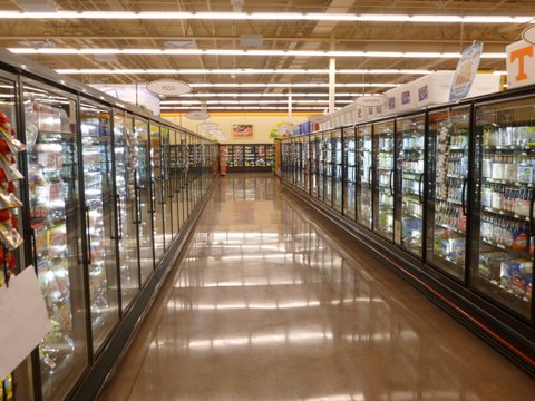 An earth-toned, polished concrete floor aisle runs between two rows of glass door refrigerators at a grocery store.
